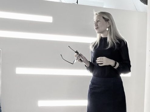 Frau mit blonden Haaren hält Brille und spricht vor einer hellen Wand.