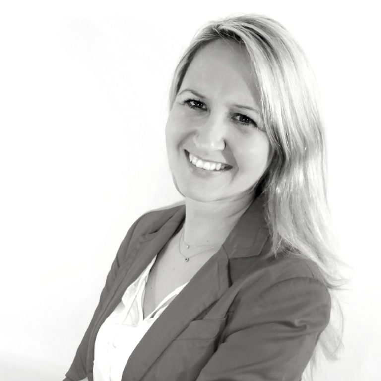 Frau mit langen blonden Haaren und Lächeln, in einem weißen Oberteil und grauem Blazer.